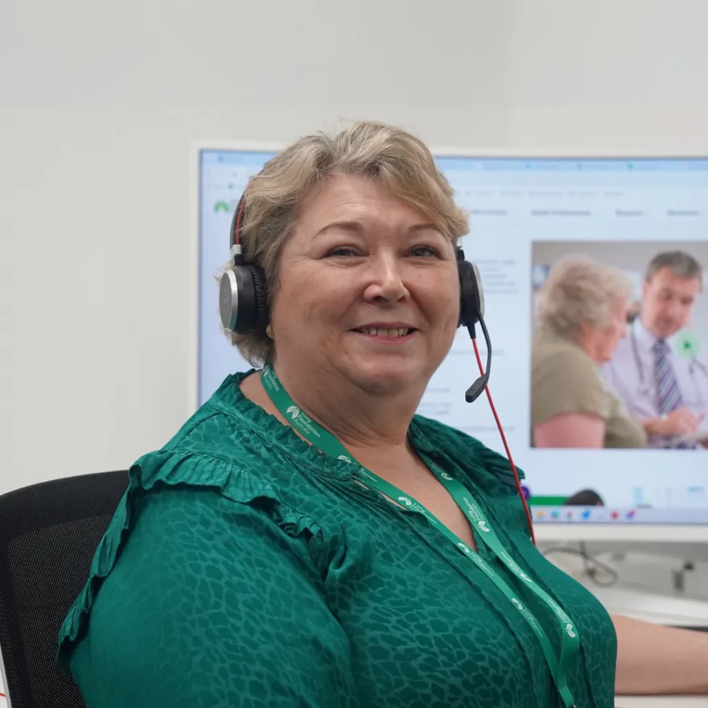 Woman sits at desk facing away from computer looking directly at camera and wearing a telephone headset.